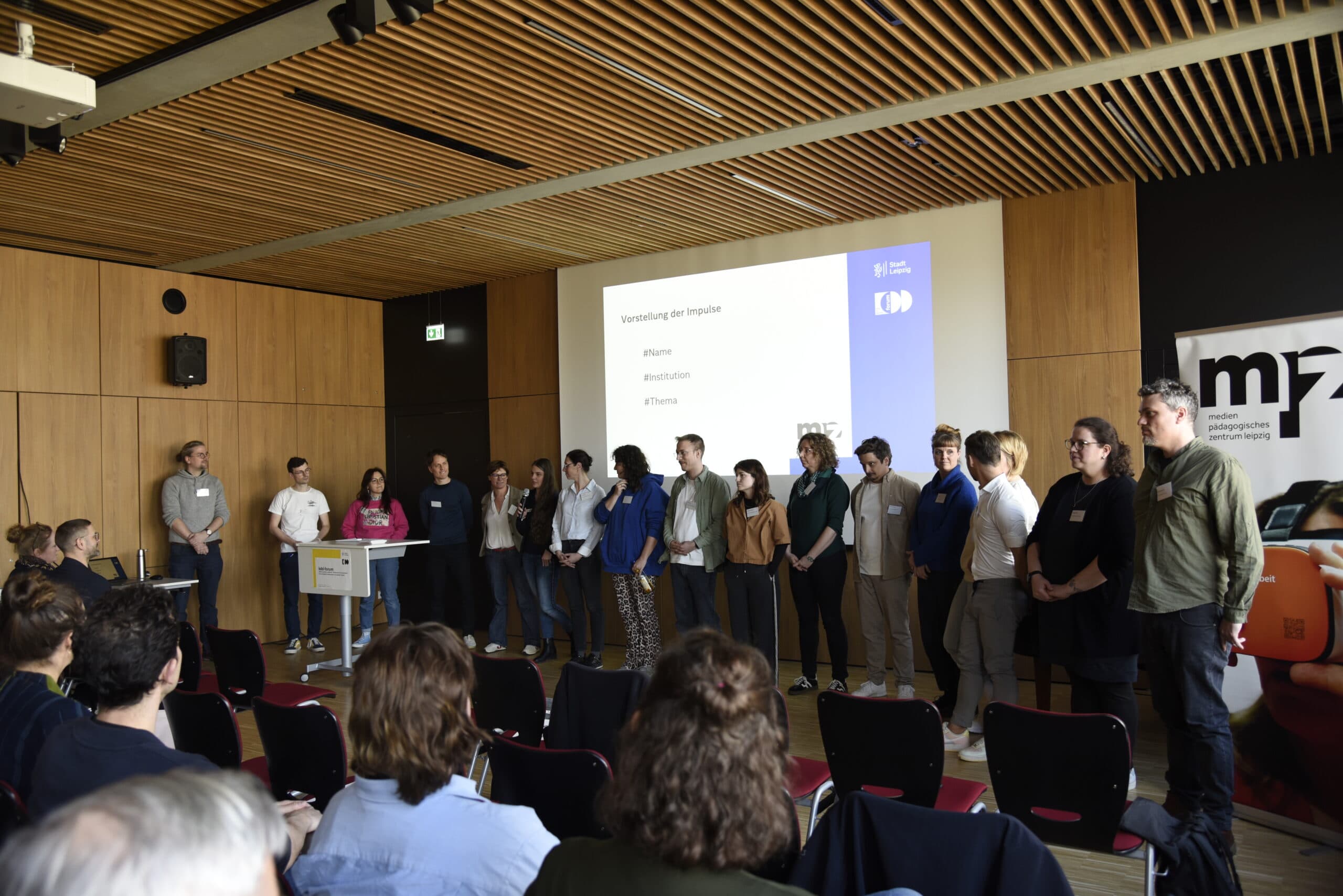 Gruppe von Personen steht vor Publikum in einem Raum mit Holzpaneelen und einer Projektionsfläche mit Text zur Vorstellung von Impulsen