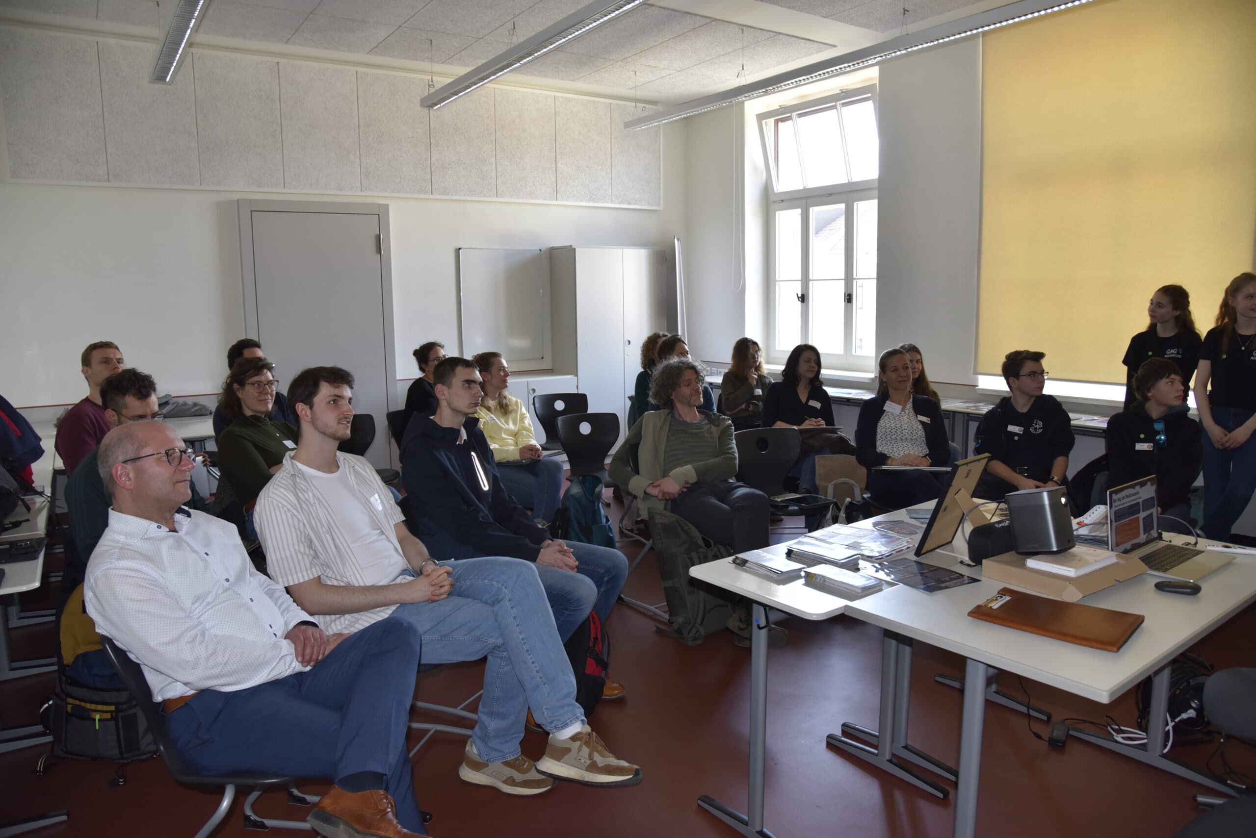 Workshops - Gruppe von Personen sitzt in einem hellen Seminarraum mit großen Fenstern und blickt auf einen Tisch mit Laptops und Präsentationsmaterial