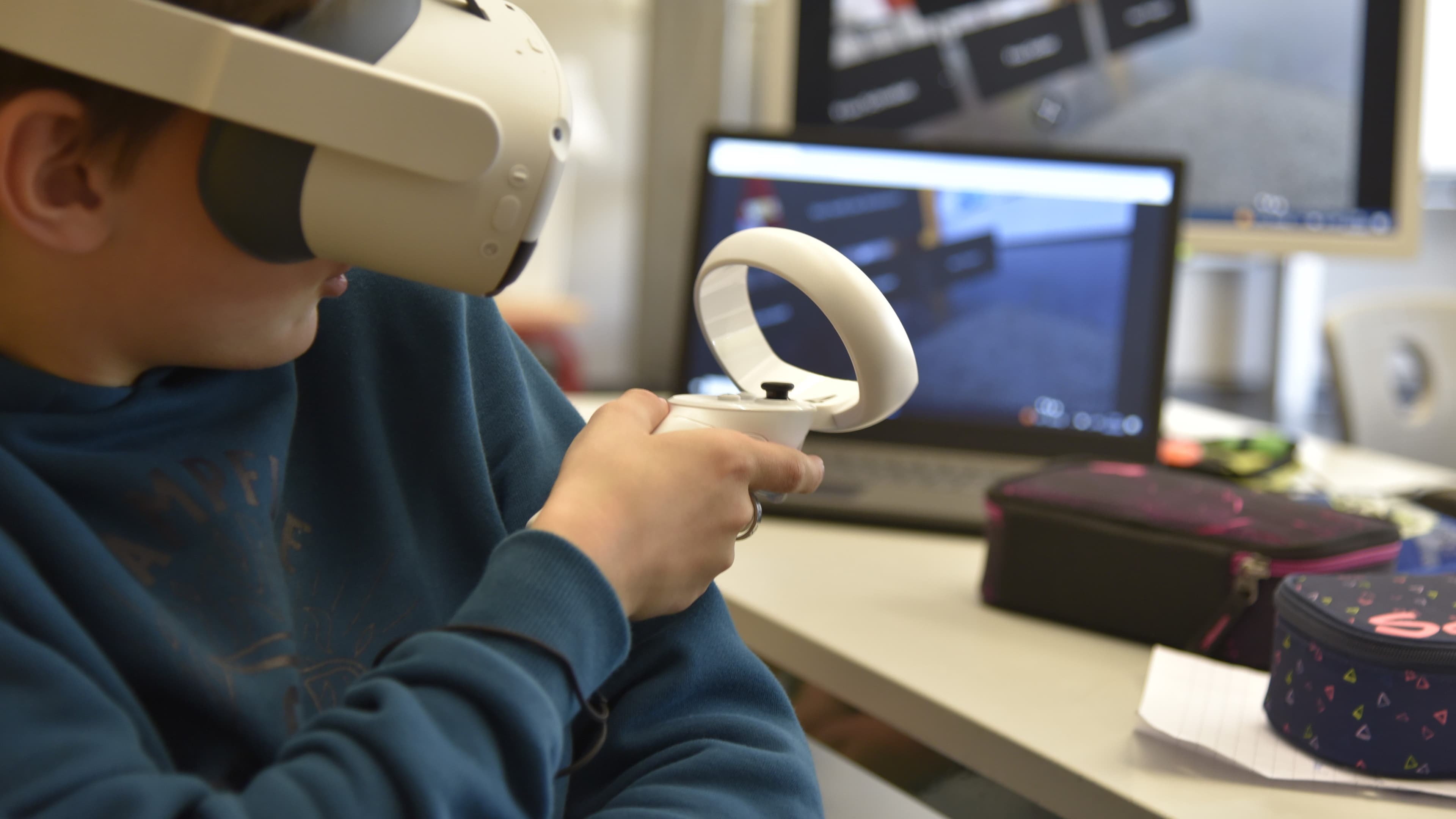 Eine Person sitzt mit einer VR-Brille auf dem Kopf und einem Controller in der Hand vor einem Bildschirm